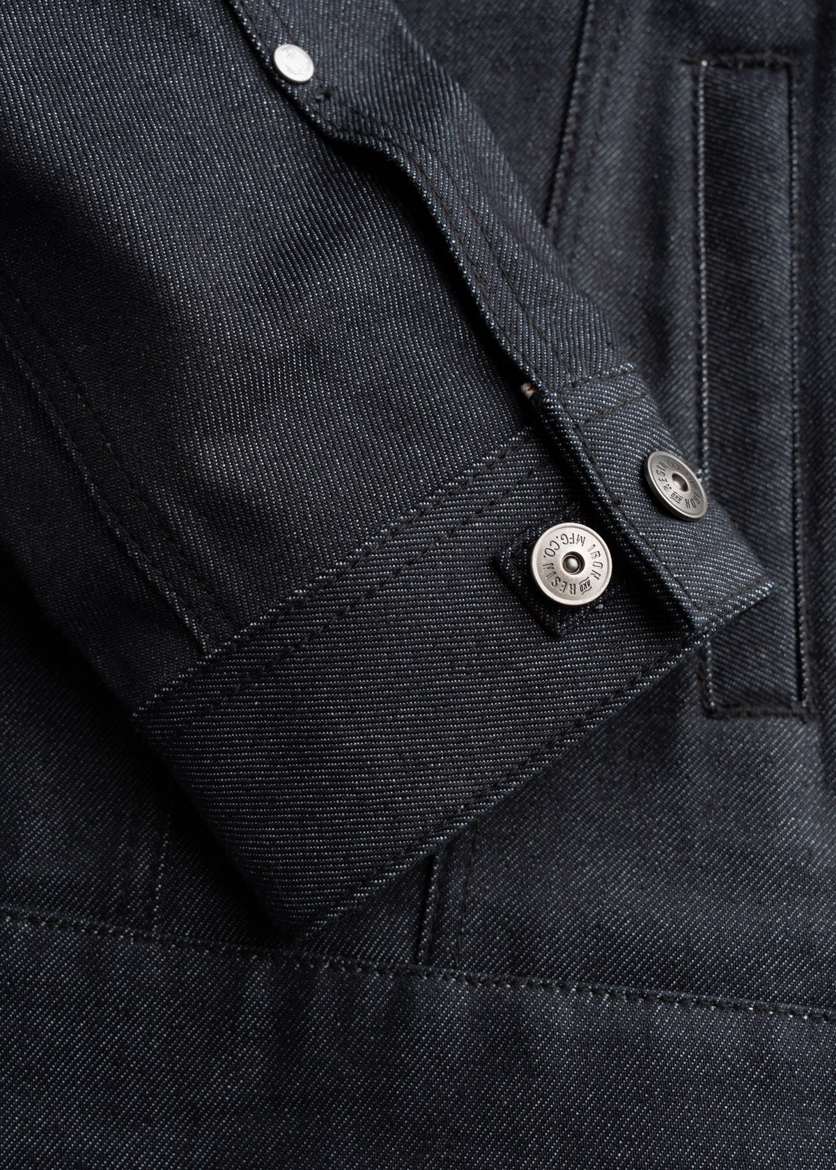 Close-up of a dark denim jacket sleeve with visible stitching and metal button details, showcasing texture and craftsmanship.