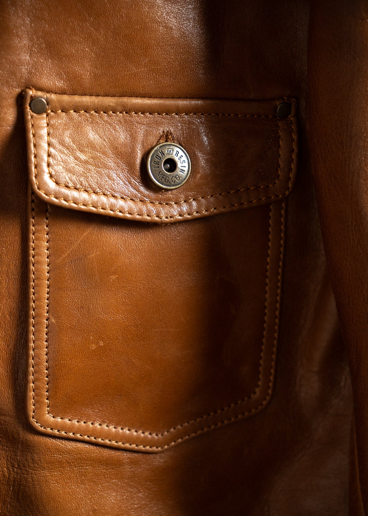 Close-up of a brown leather jacket pocket with a metal button engraved "Iron Resin MFG Co," showcasing detailed stitching.