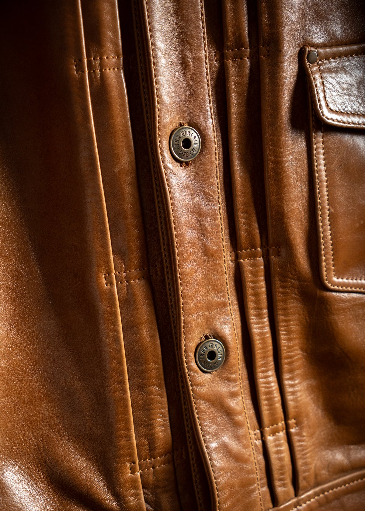 Close-up of a brown leather jacket with detailed stitching and metal buttons, showcasing texture and craftsmanship.