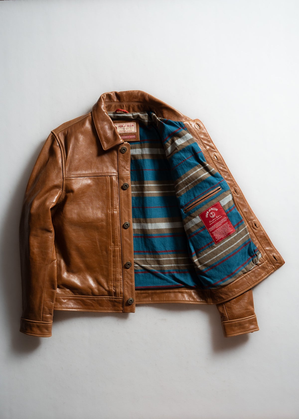 Brown leather jacket with open front reveals blue and brown striped lining. Red label inside. Laid on a white background.