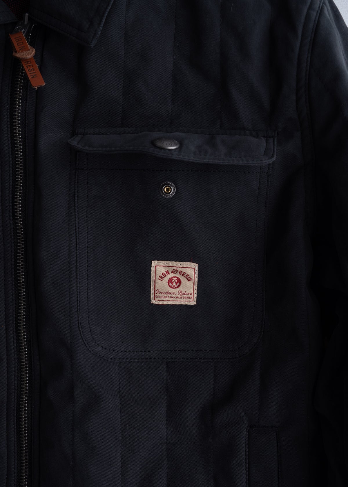 Close-up of a black jacket with a front pocket, featuring an "Iron and Resin" label. A brown zipper pull is visible.