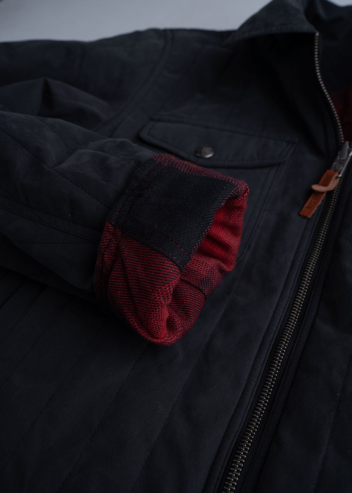Black jacket with red plaid lining, zipper, and buttoned pocket. The sleeve is folded, revealing the inner fabric.