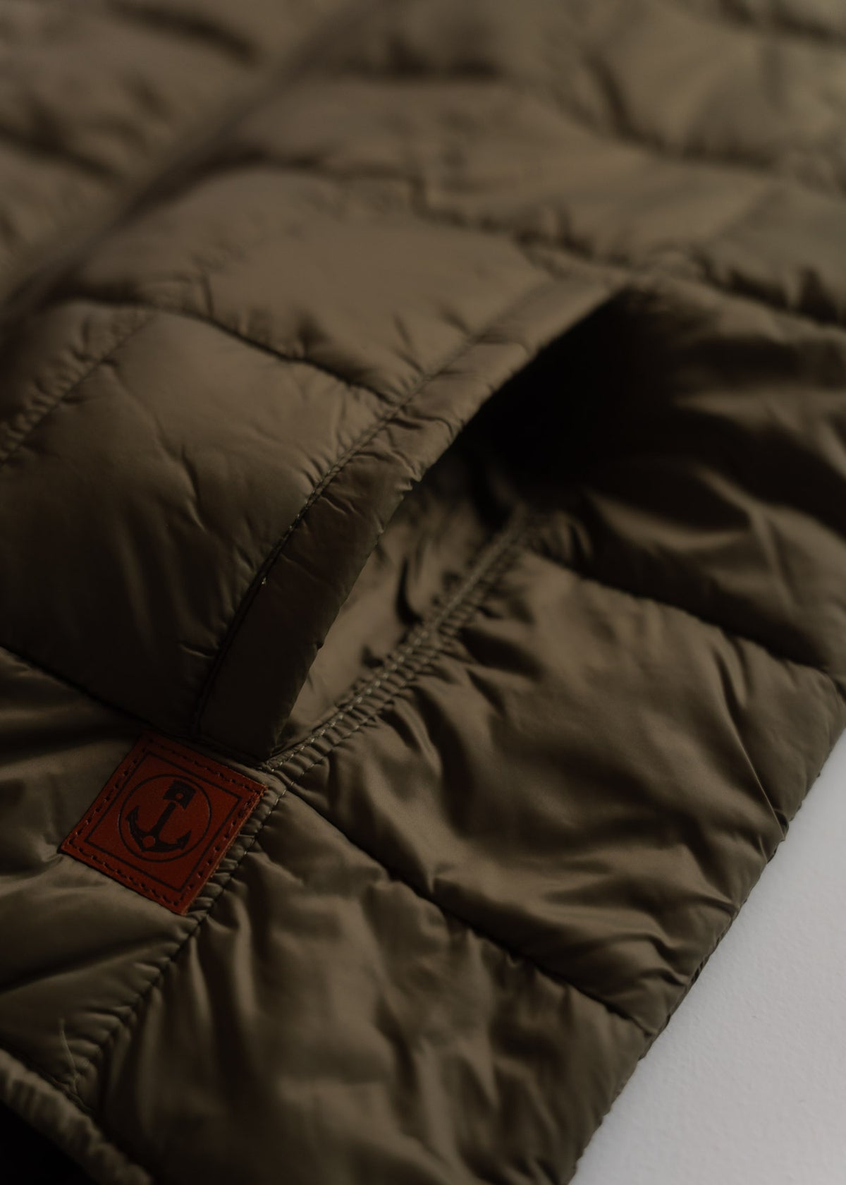 Close-up of a brown quilted jacket with a visible pocket and anchor logo patch on a white surface, highlighting texture.