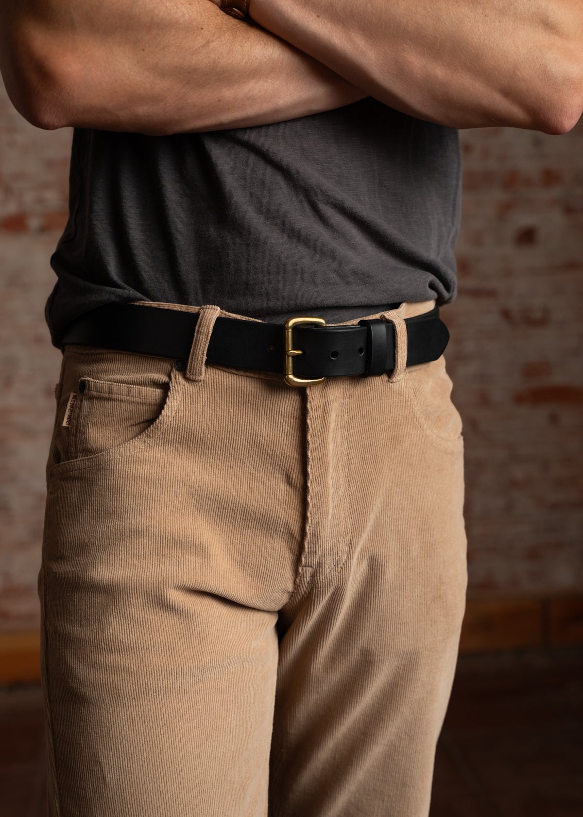 Man in a gray shirt and tan corduroy pants with a black belt stands against a brick wall, arms crossed confidently.
