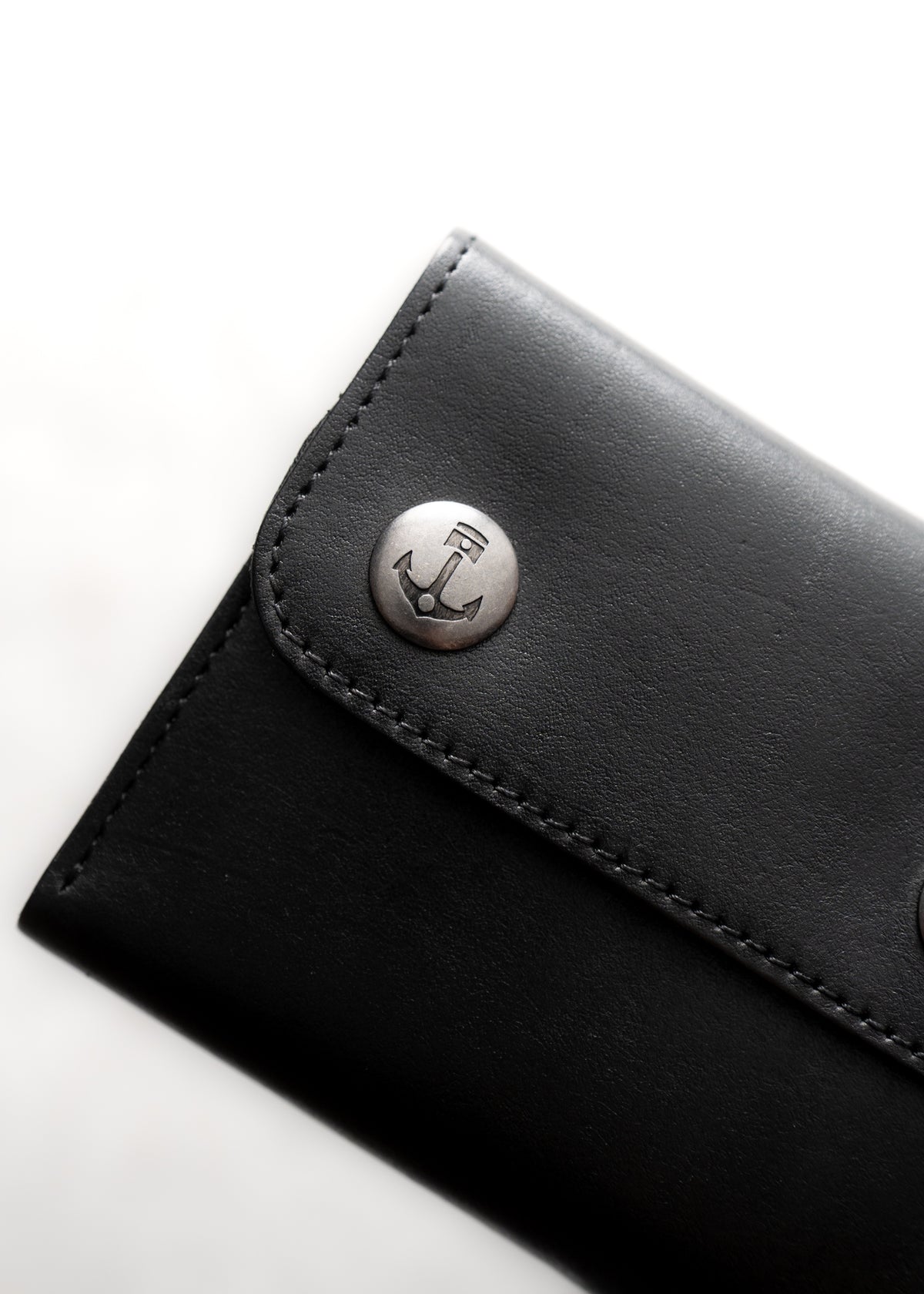 Black leather wallet with metal button featuring an engraved anchor design, highlighting a nautical theme against a white background.
