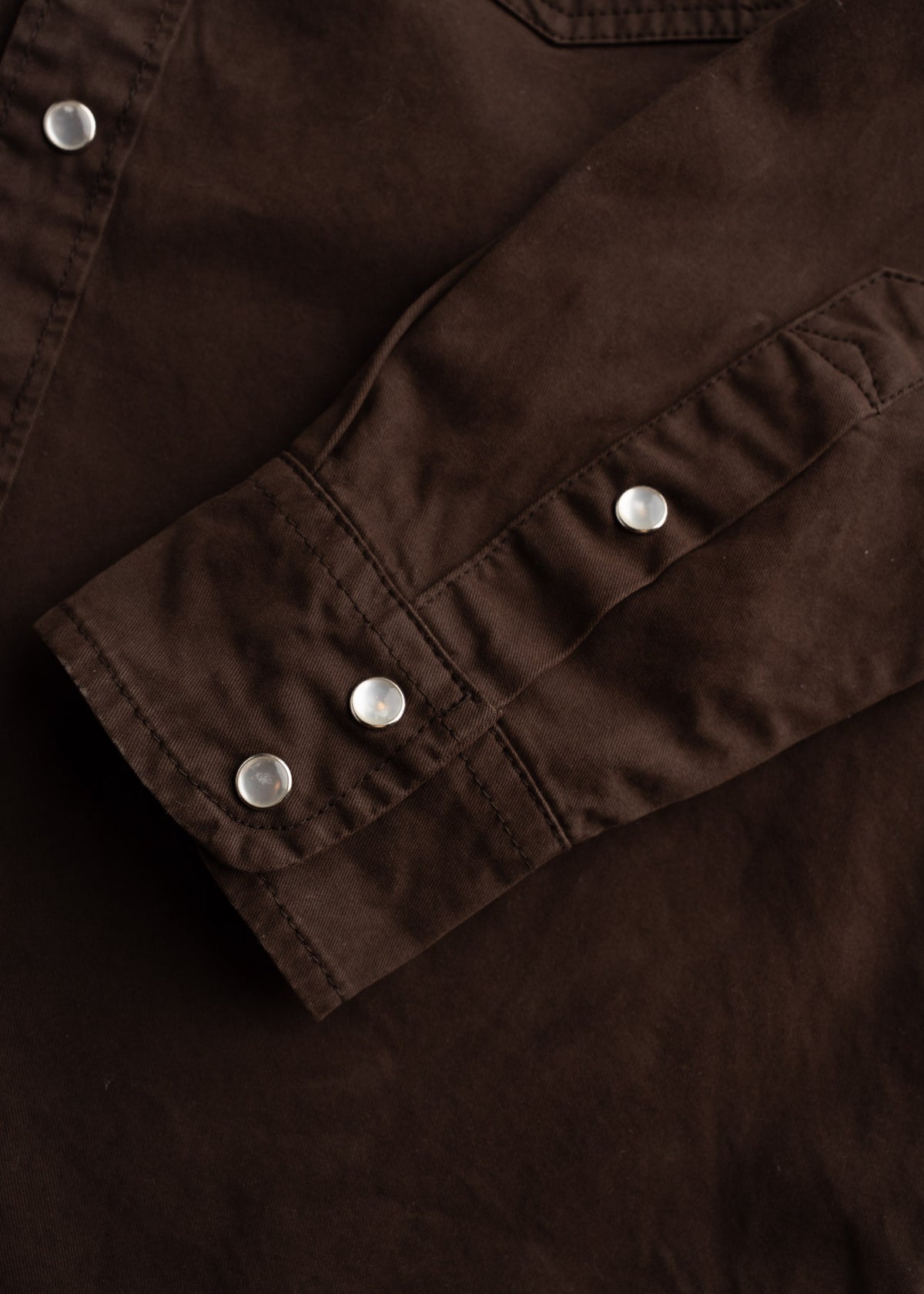 Close-up of a brown shirt sleeve with silver snap buttons, showcasing detailed stitching and fabric texture.
