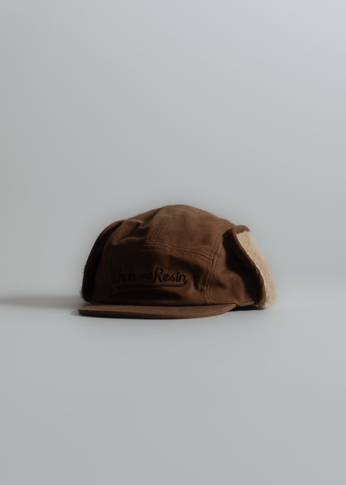 Brown cap with ear flaps, labeled "Iron and Resin," against a light gray background. Cozy winter accessory.