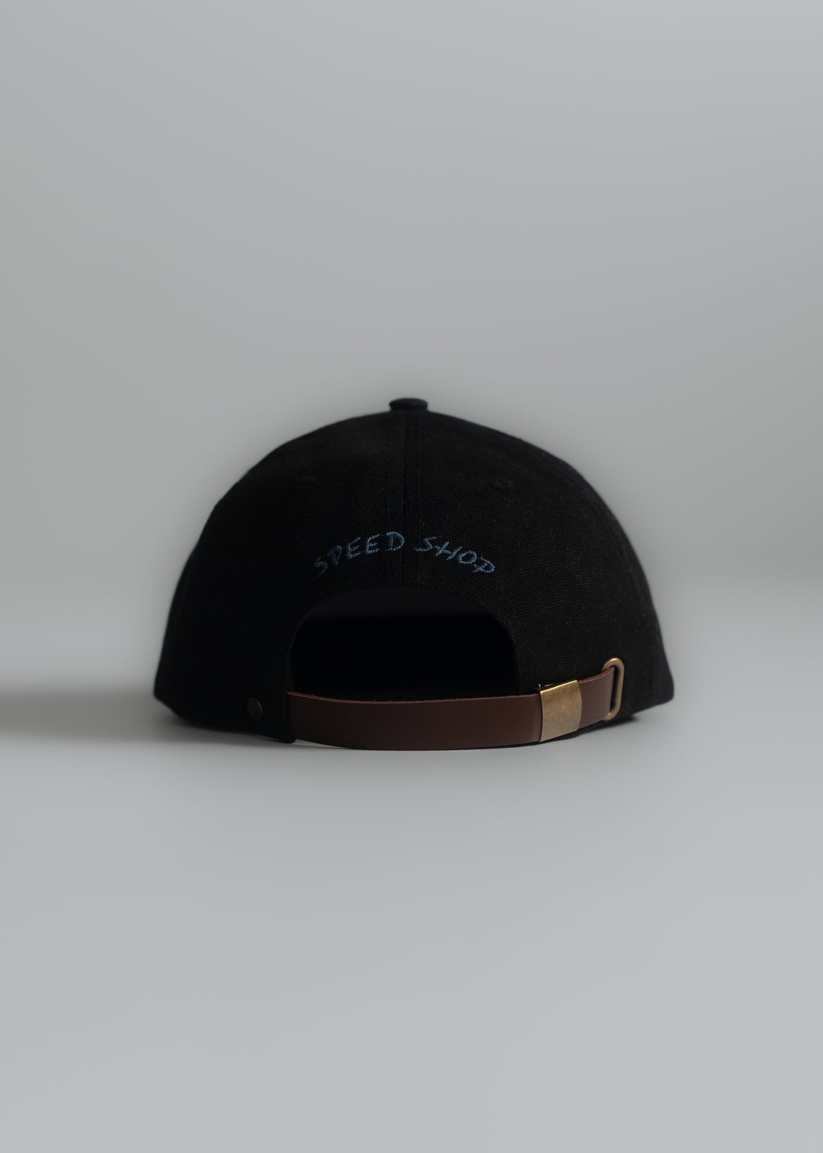 Black cap with "Speed Shop" text, brown adjustable strap, and gold buckle on a light gray background, viewed from the back.
