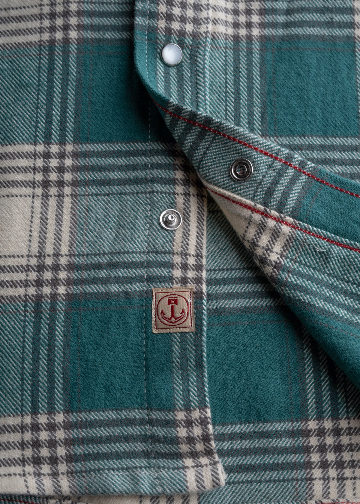 Close-up of a teal plaid shirt with snap buttons and red stitching, featuring an anchor logo patch on the fabric.