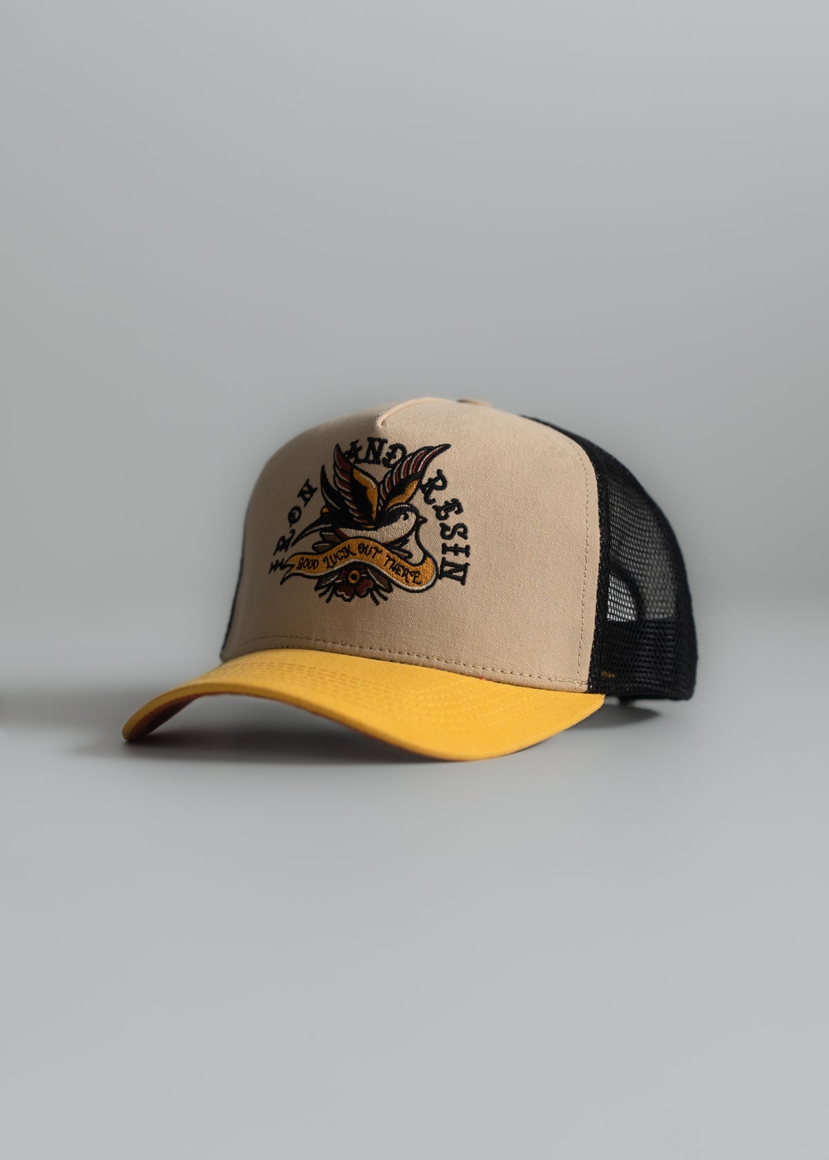 A beige cap with a yellow brim features an embroidered eagle design and black mesh back, set against a neutral background.