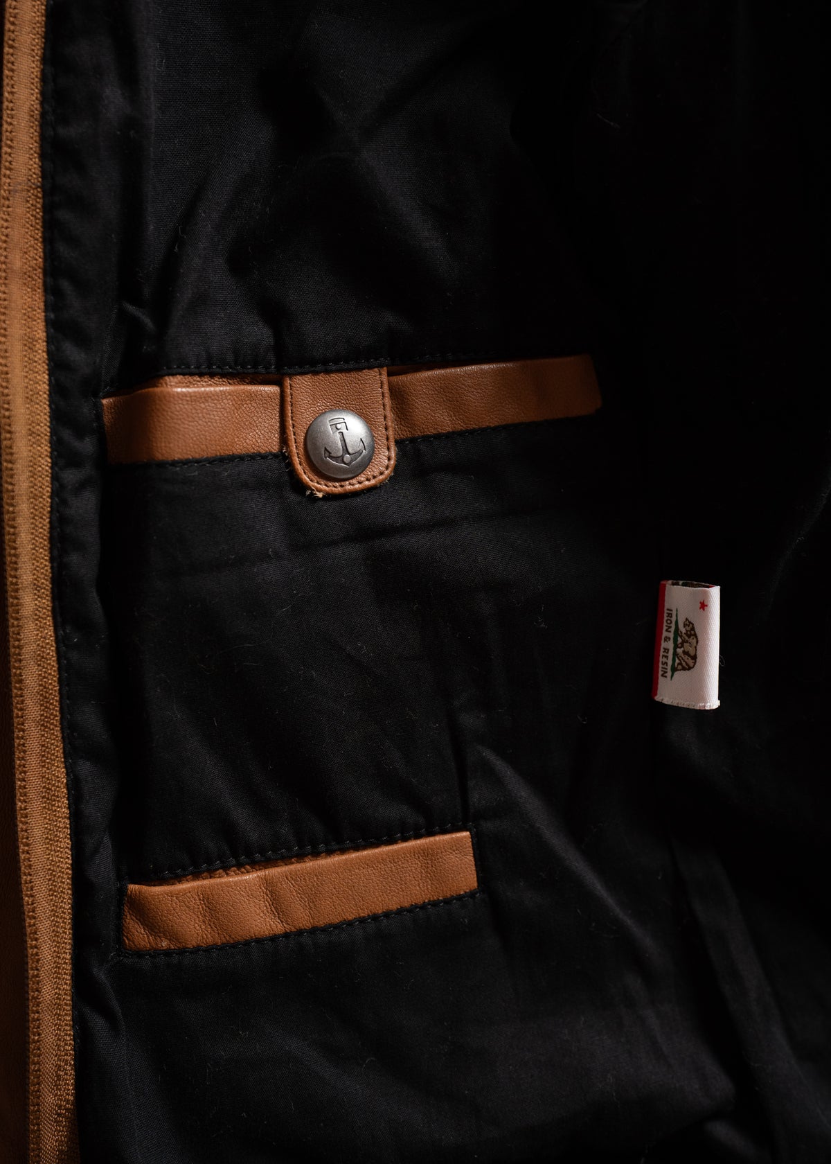 Black jacket interior with brown leather trim, anchor button, and a small white label. Simple and stylish design elements.