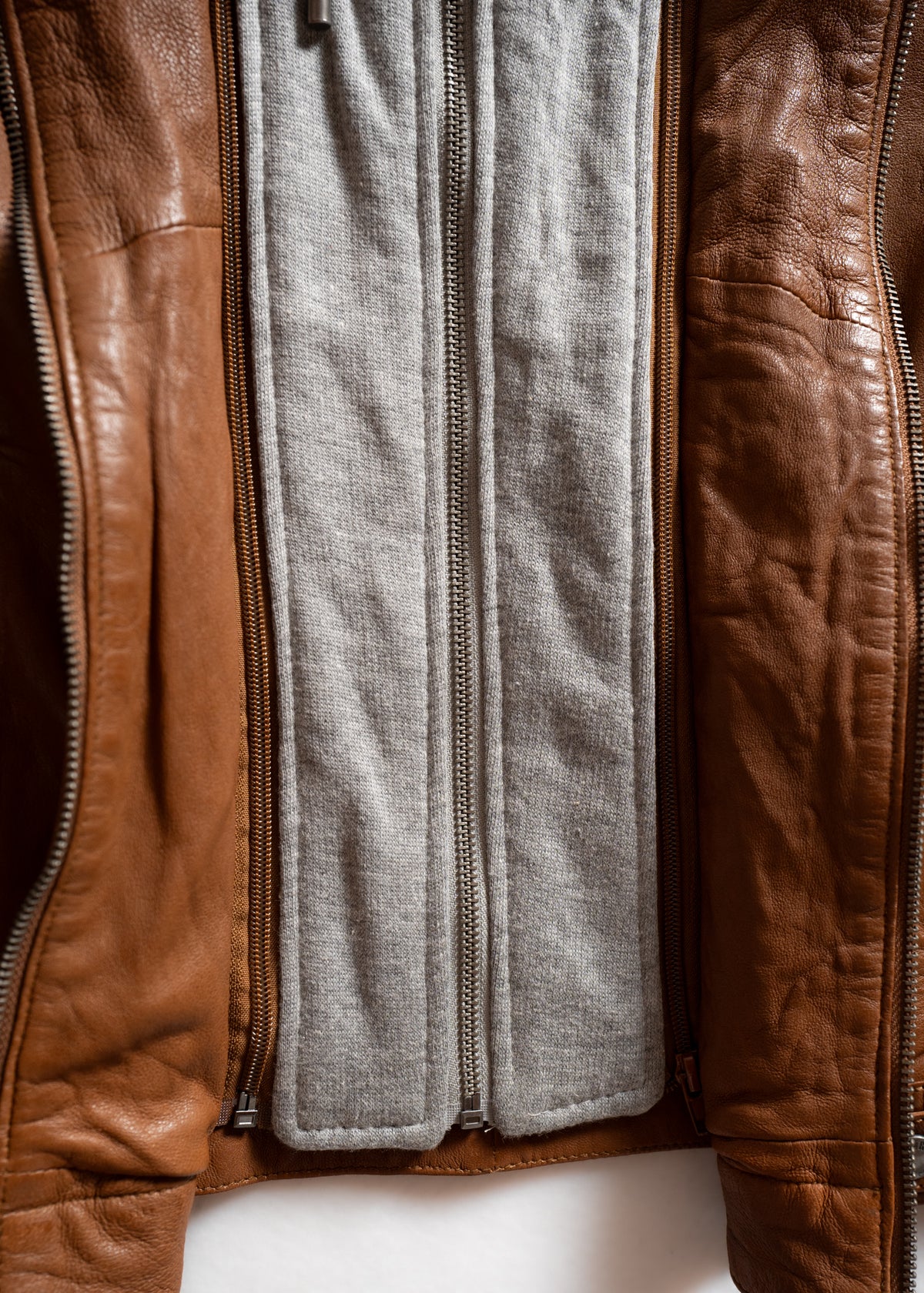 Open brown leather jacket revealing a gray fabric lining with a central zipper, highlighting texture and design details.