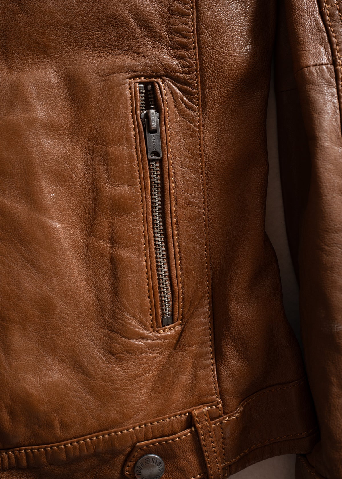 Close-up of a brown leather jacket showing a vertical zippered pocket and button detail, highlighting texture and craftsmanship.