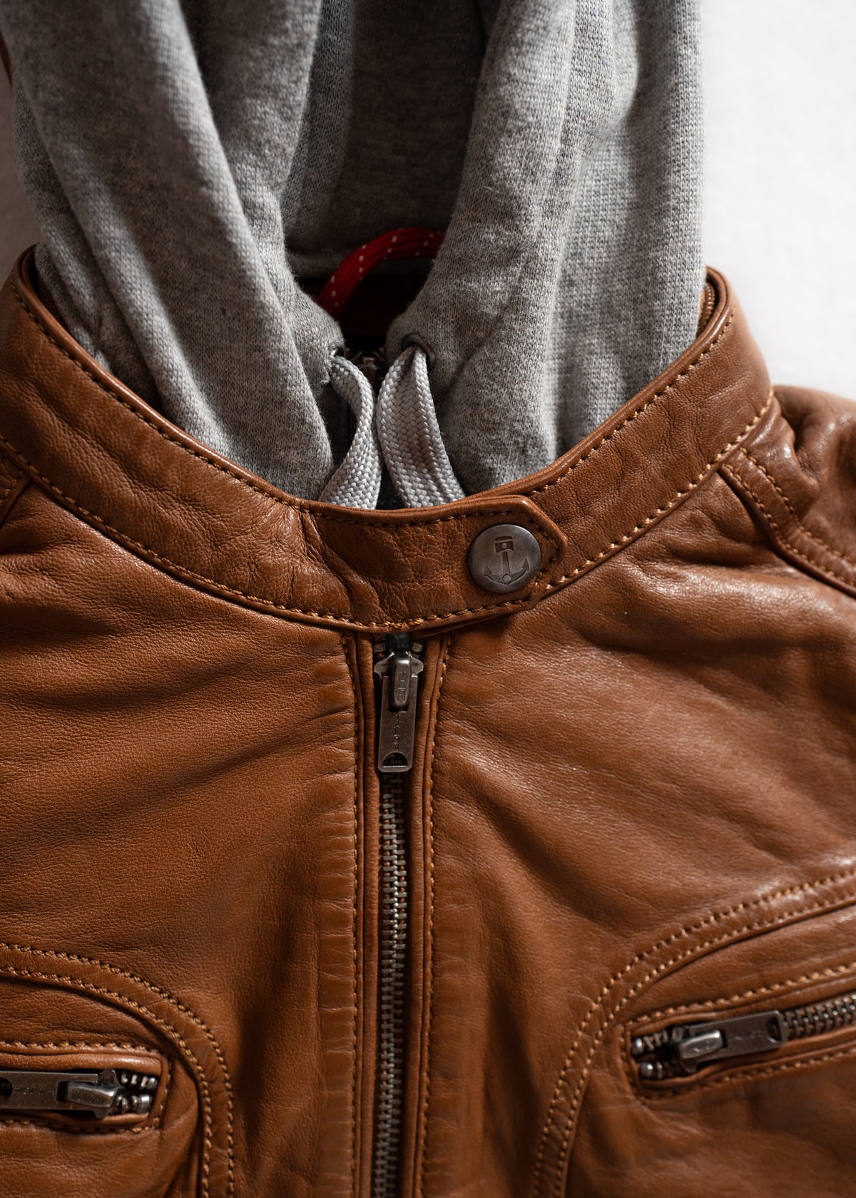 Brown leather jacket with a gray hoodie underneath, featuring zippers and a button detail, showcasing a casual layered style.