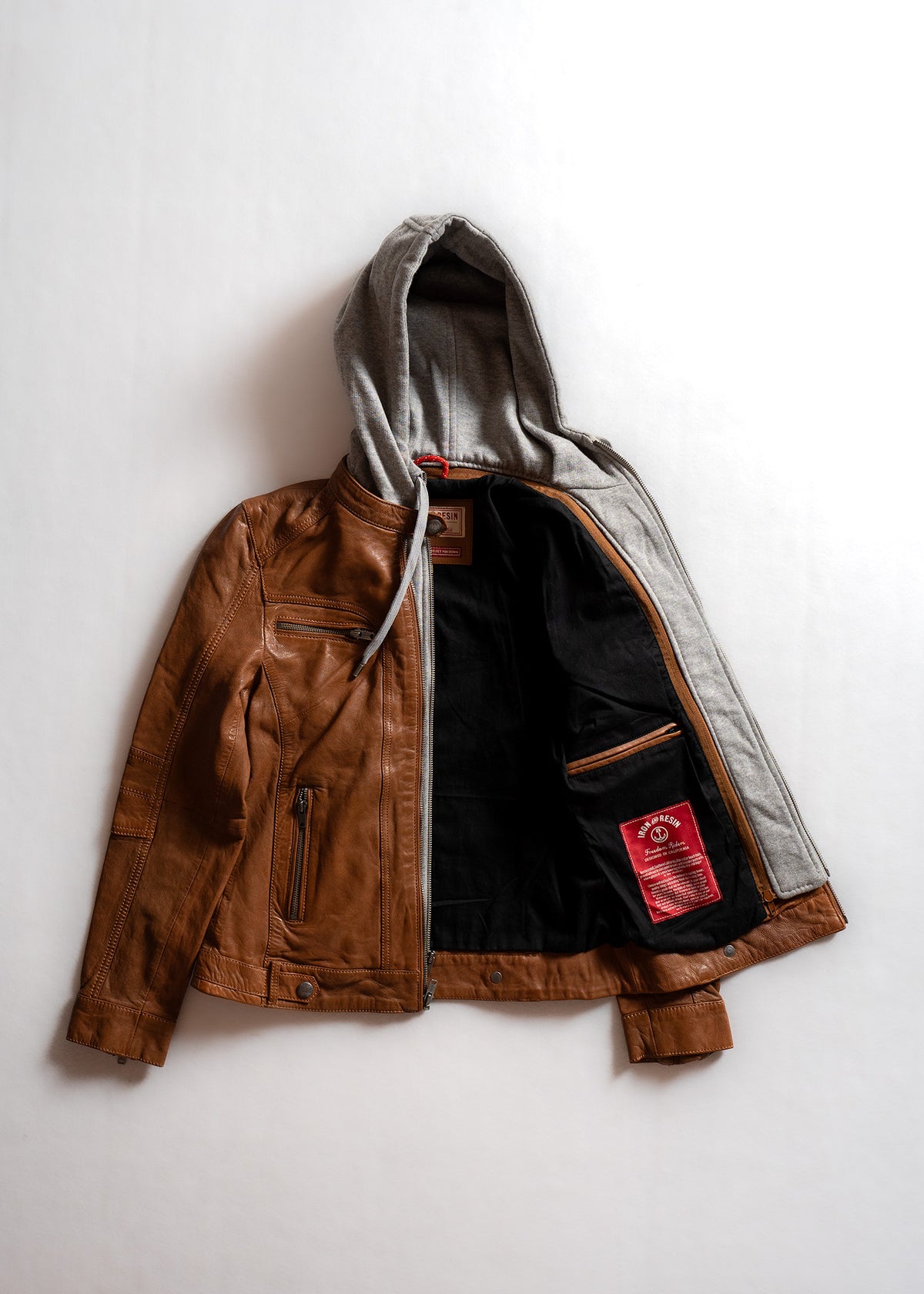 Brown leather jacket with a gray hood, open to reveal a black interior and red label, lying flat on a white background.