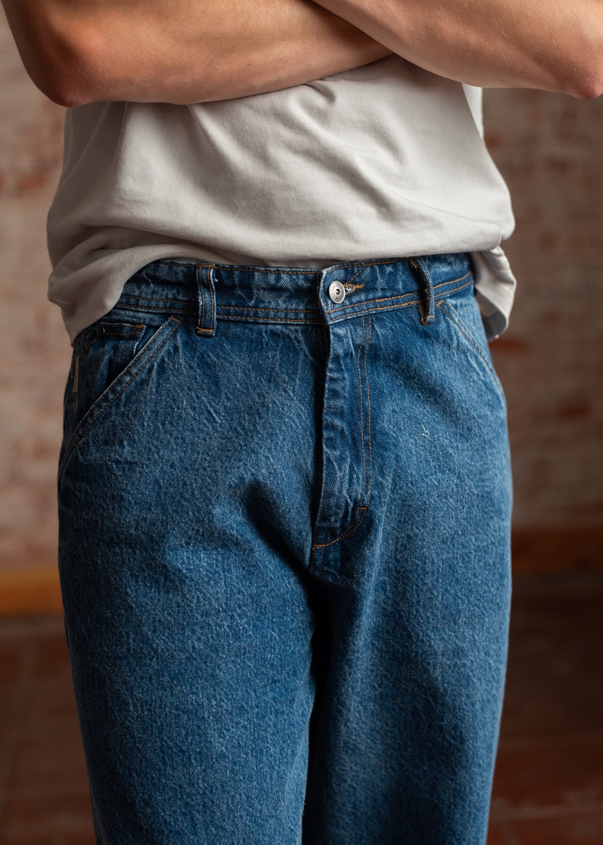 Person in blue jeans and a white shirt stands with arms crossed, showing casual style against a blurred brick background.