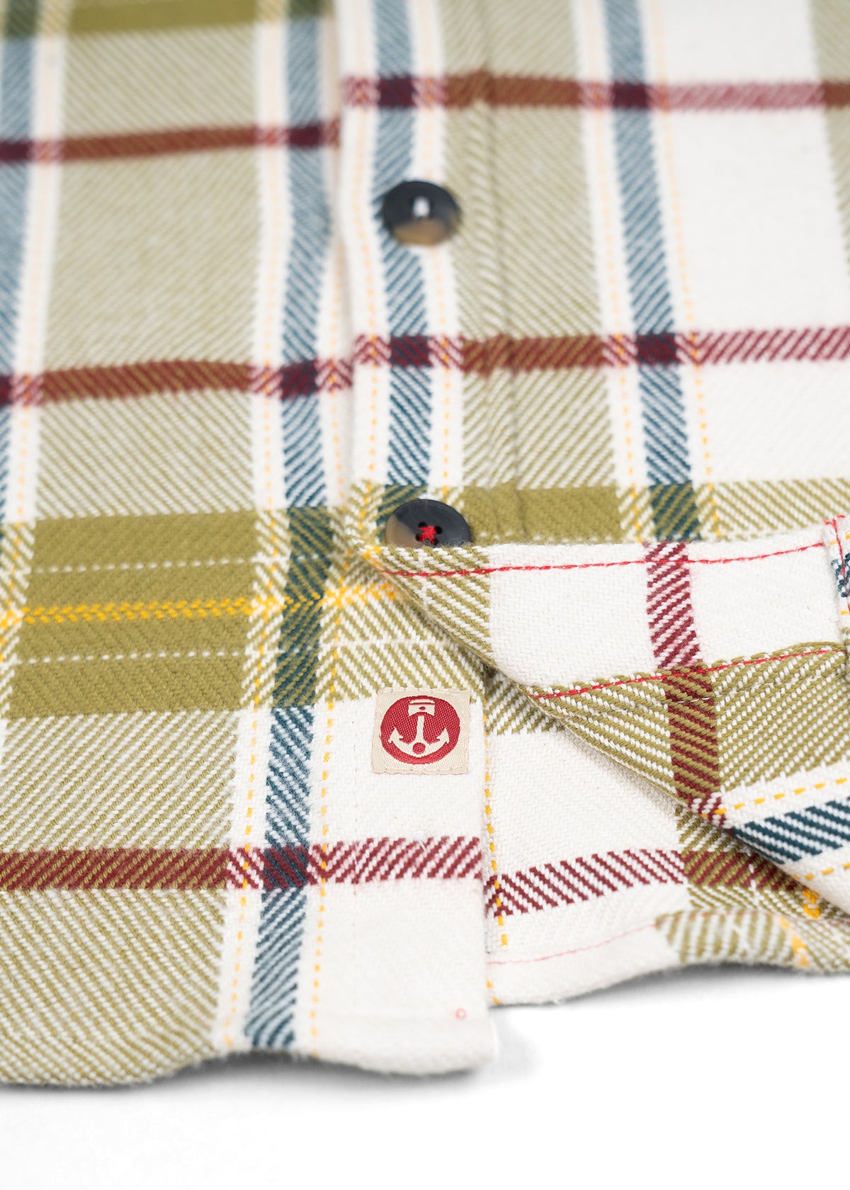Close-up of a plaid shirt with green, red, and blue stripes, featuring black buttons and an anchor logo on the fabric.