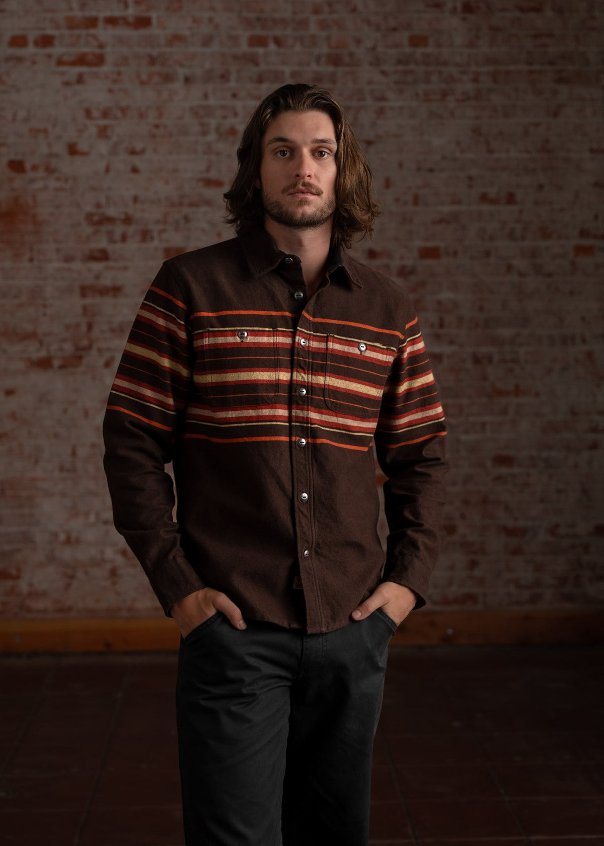 Man in a brown striped shirt stands confidently against a rustic brick wall, hands in pockets, exuding a relaxed, stylish vibe.