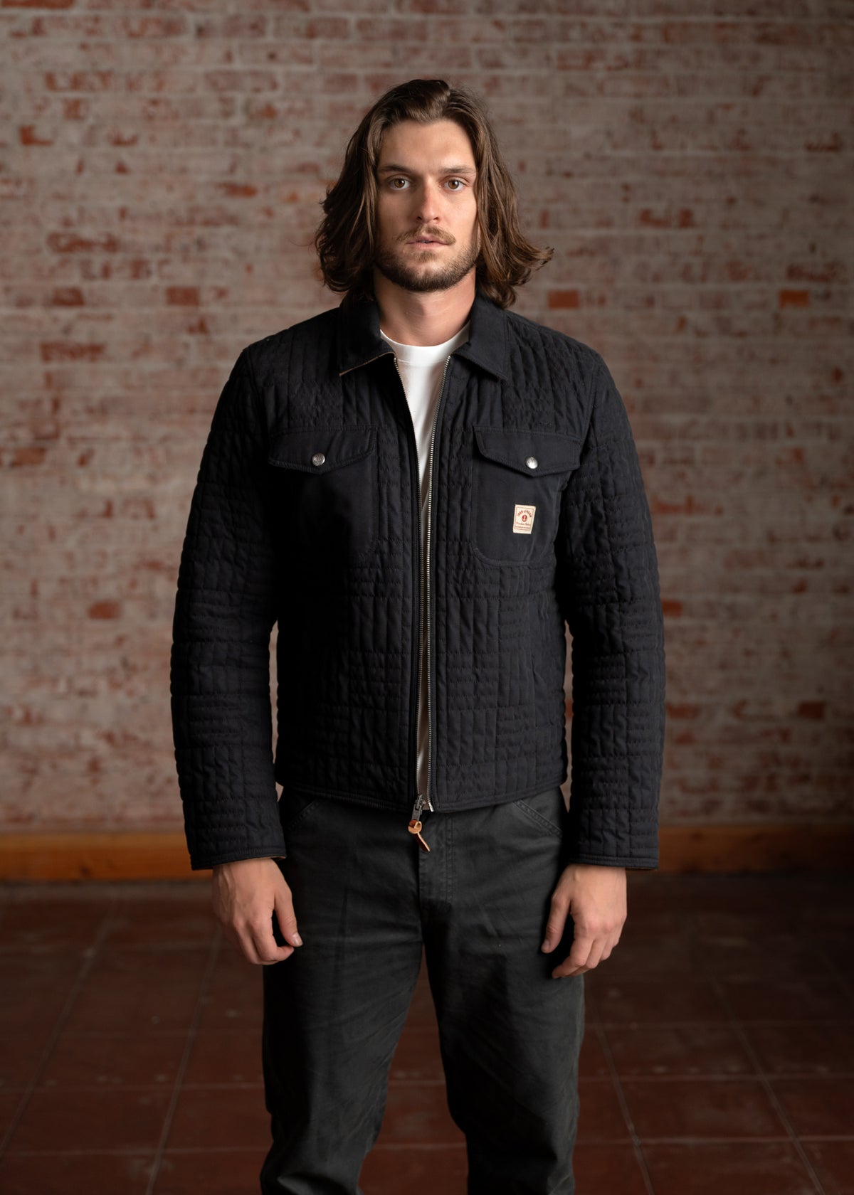 Man with long hair in a black quilted jacket stands against a brick wall, looking directly at the camera with a serious expression.