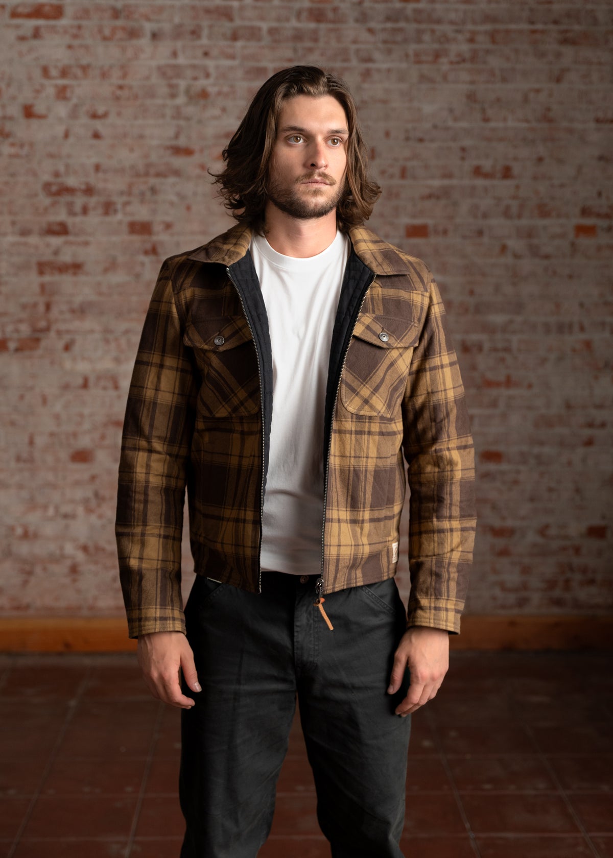Man in a plaid jacket and white shirt stands against a brick wall, looking forward. Casual, stylish, and confident pose.