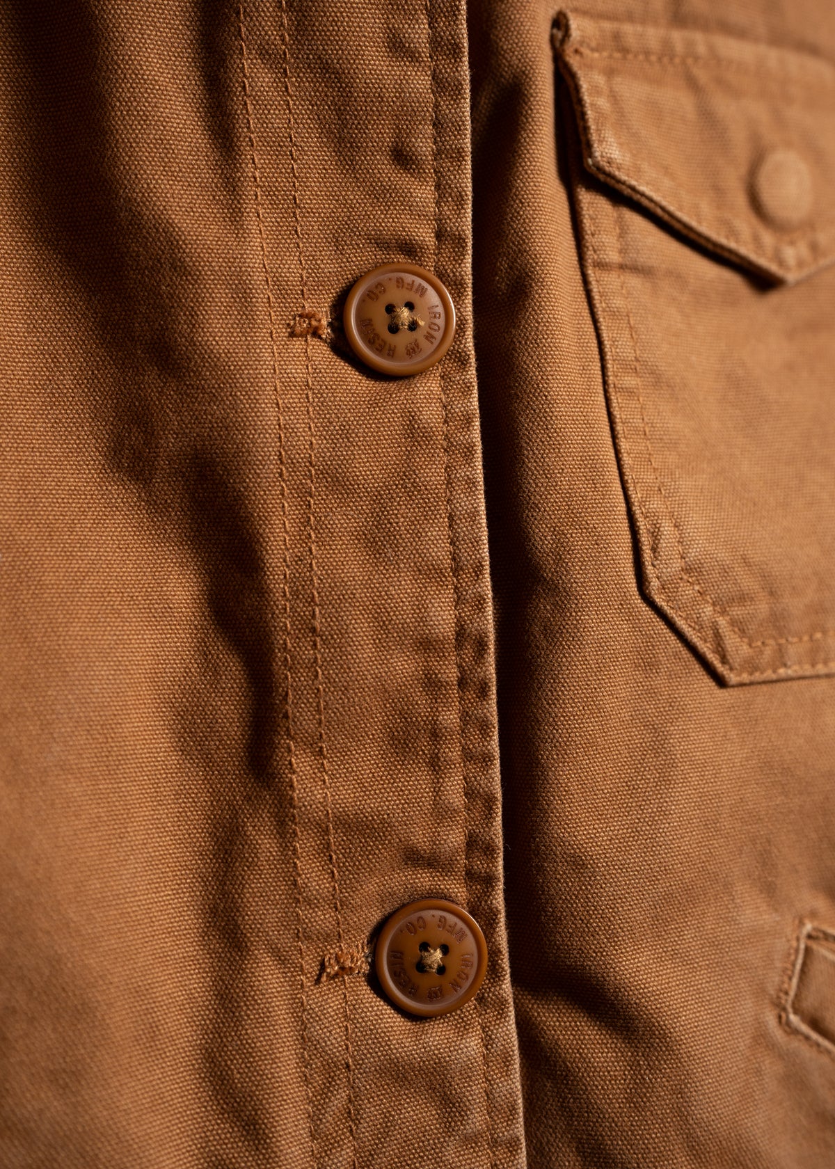Close-up of a brown shirt showing two buttons and a chest pocket, highlighting the fabric texture and stitching details.
