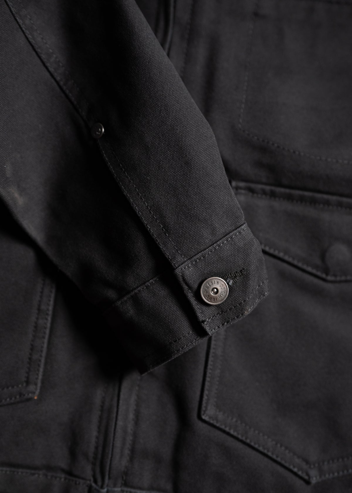 Close-up of a black denim jacket sleeve with a buttoned cuff, highlighting the texture and stitching details.