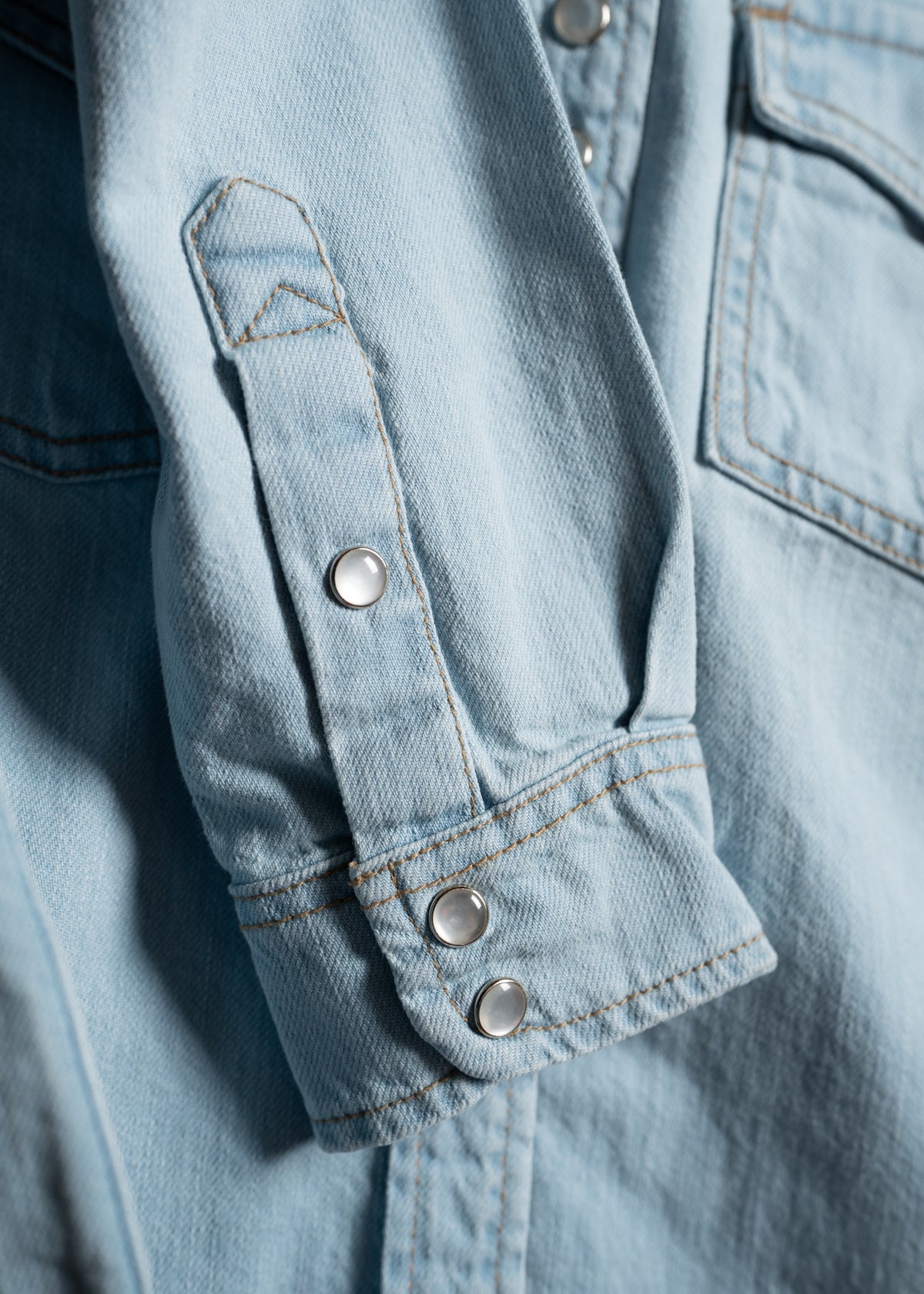 Close-up of a light blue denim shirt sleeve with silver snap buttons and visible stitching, highlighting casual fashion details.