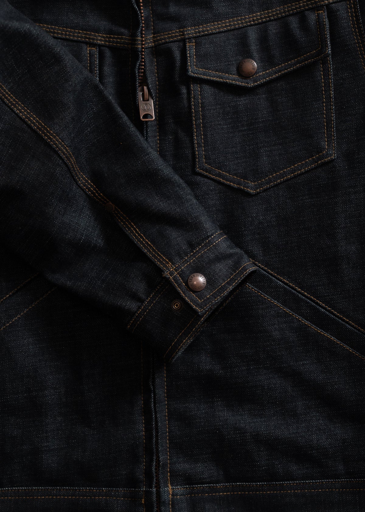 Dark denim jacket with brown stitching, featuring a front pocket, zipper, and metal buttons on the sleeve, showcasing detail.