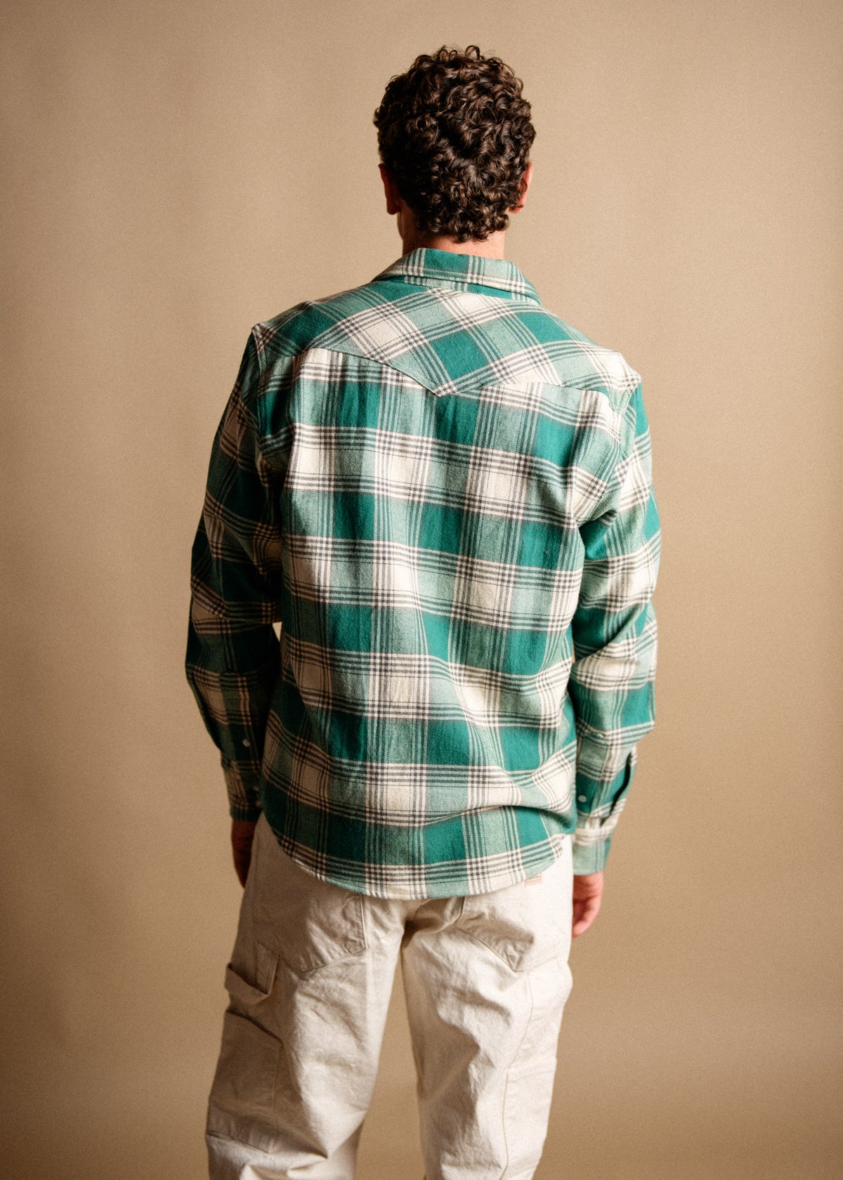 Man in green plaid shirt and beige pants stands with back to camera against a neutral background, highlighting casual style.