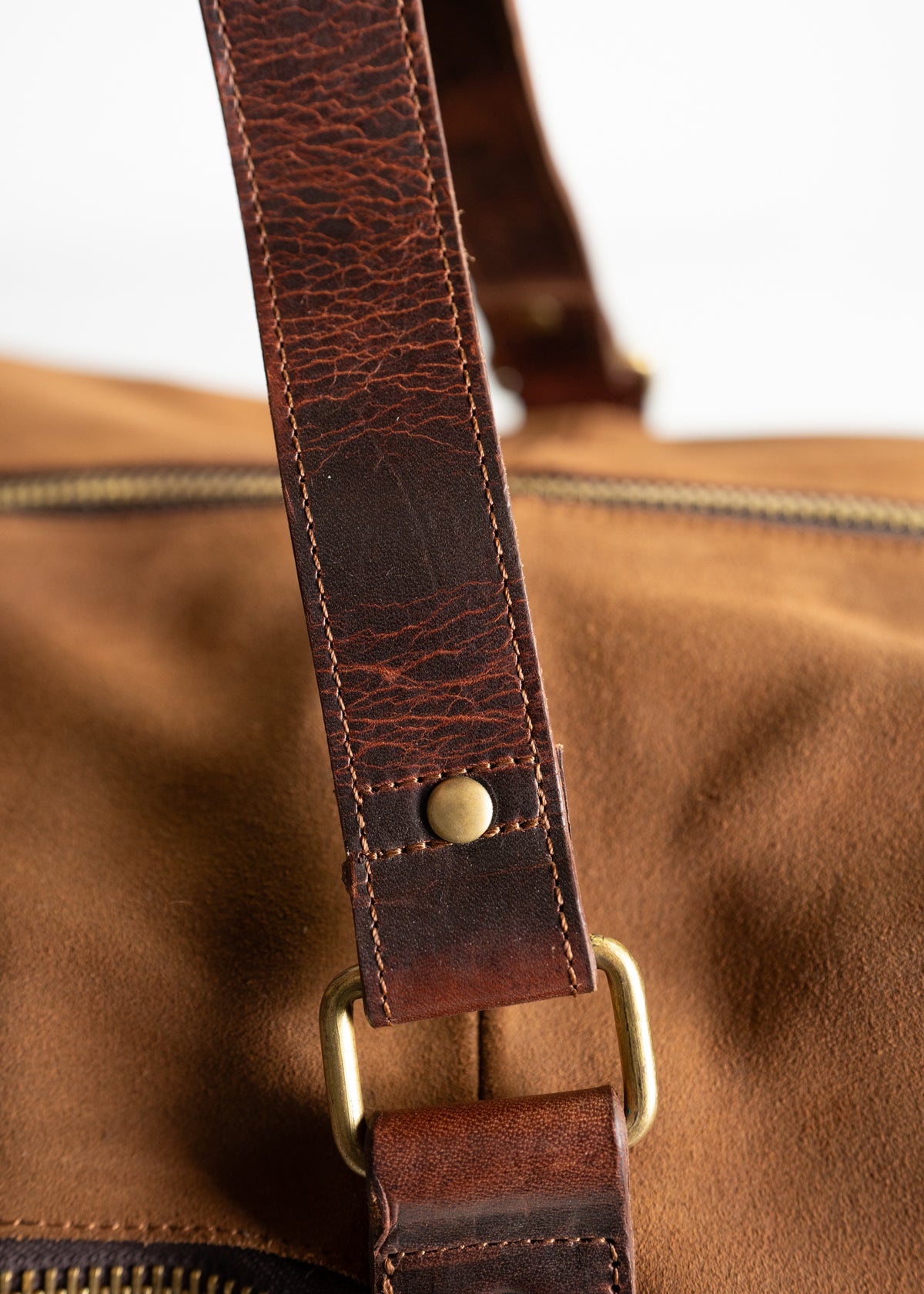 Close-up of a brown leather strap with brass hardware on a tan bag, highlighting texture and craftsmanship.