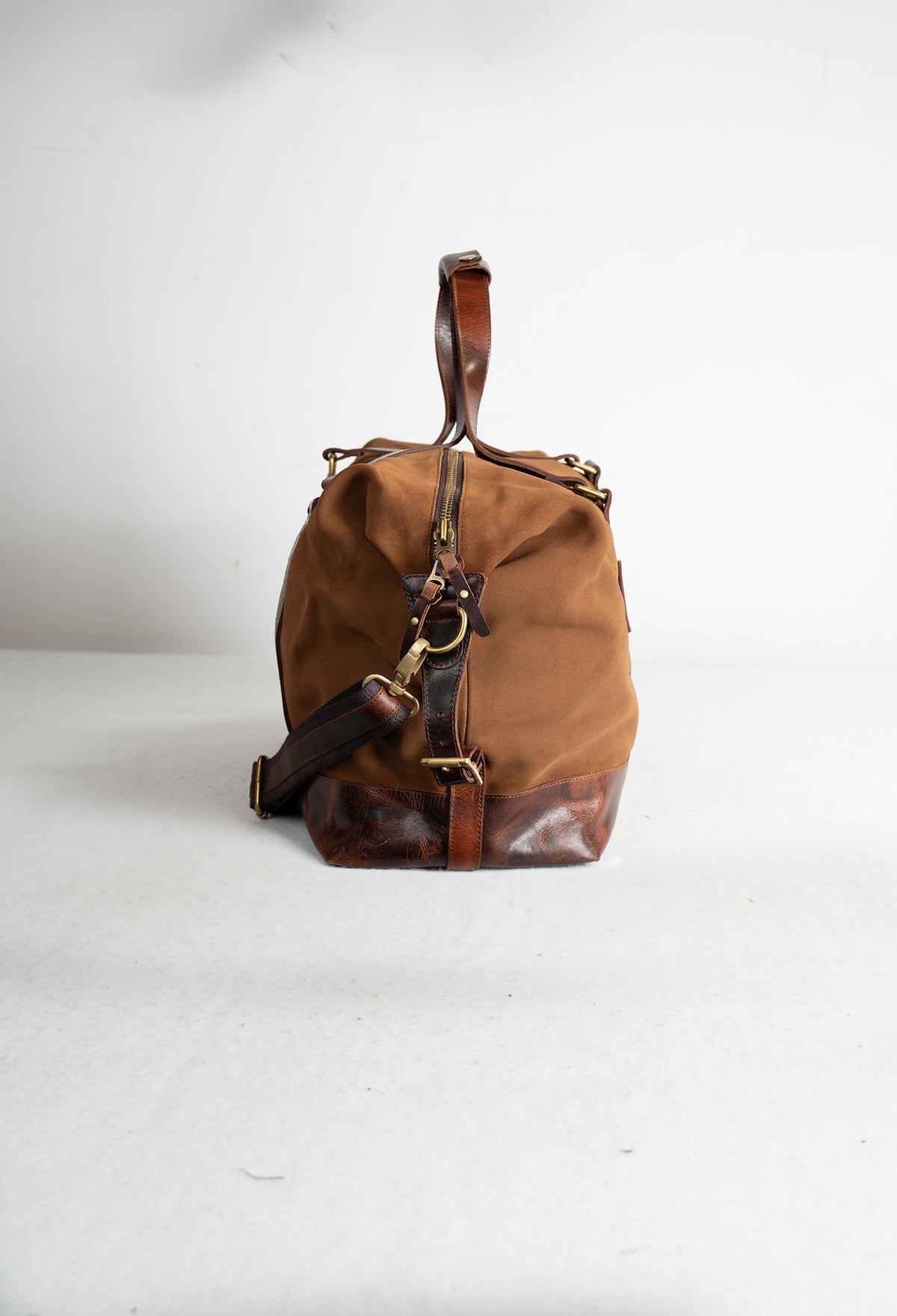 Brown canvas and leather duffle bag with a shoulder strap, standing upright on a white surface against a plain background.