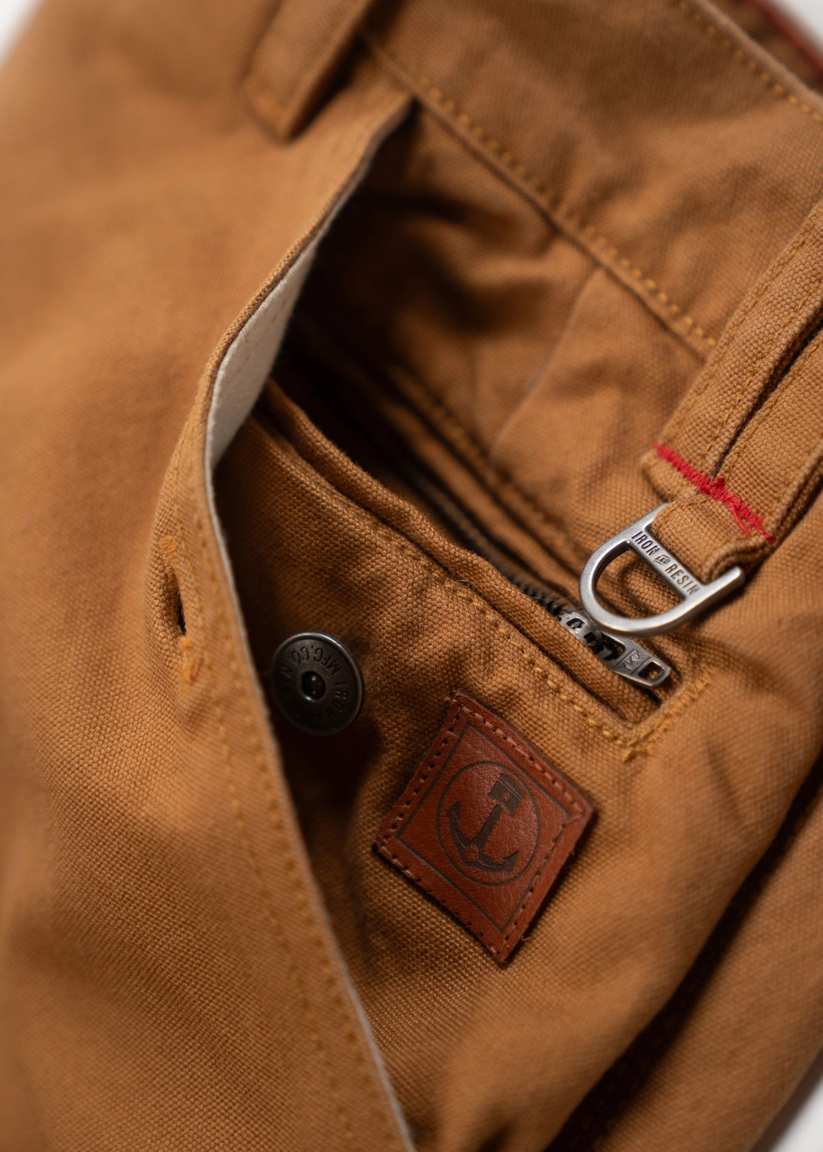 Close-up of brown pants pocket with a leather anchor patch, metal button, and zipper detail, showcasing rugged craftsmanship.