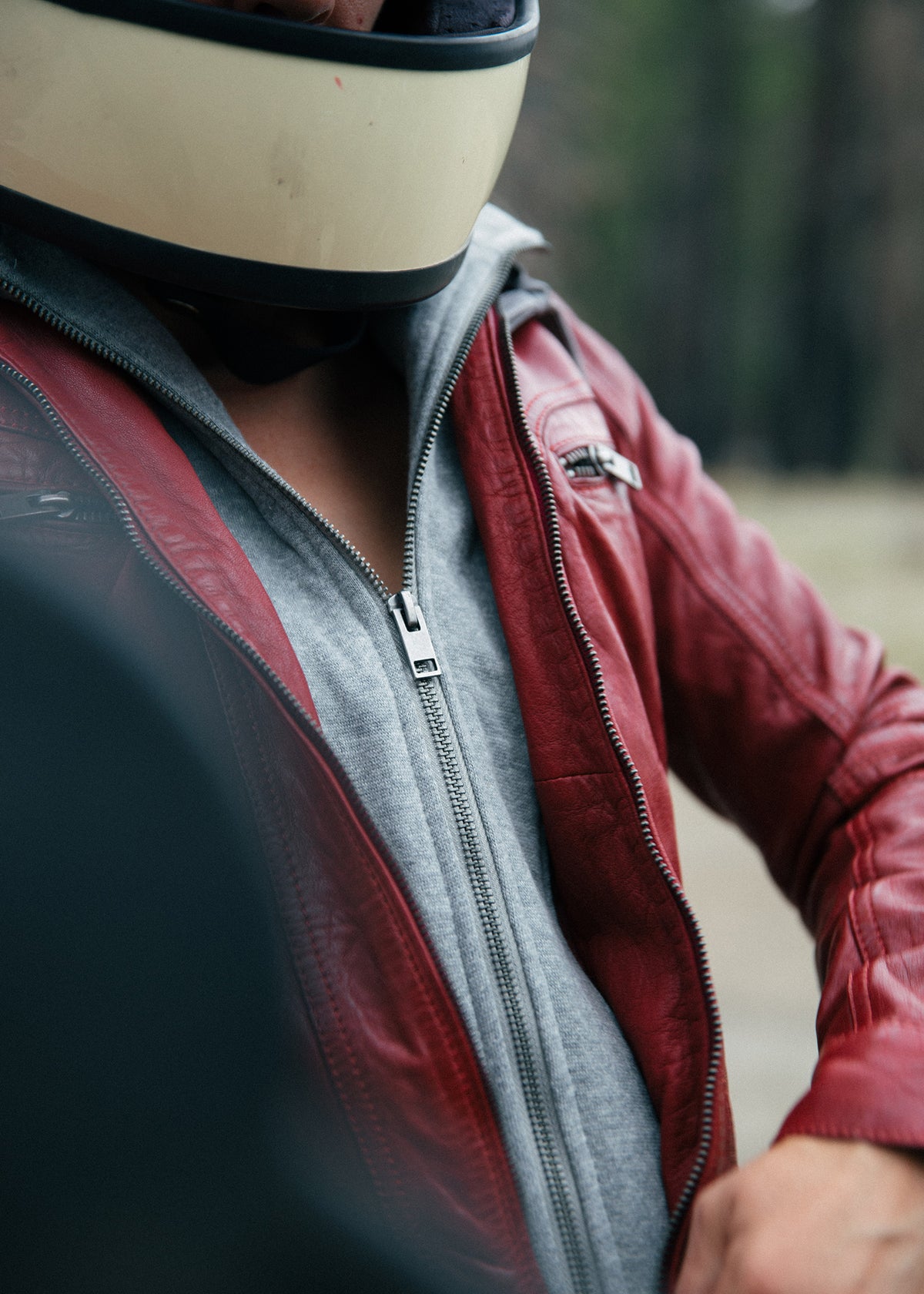 Person in a red leather jacket and helmet, partially visible, stands outdoors with blurred trees in the background.