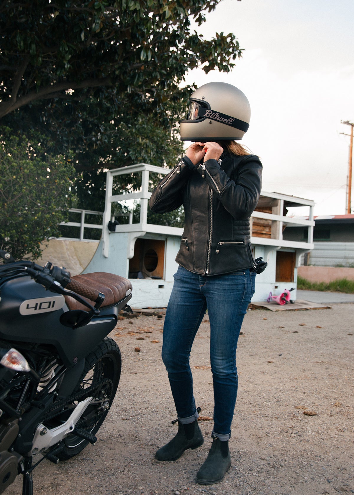 Woman in a leather jacket adjusts her helmet beside a motorcycle, ready to ride on a sunny day with trees in the background.